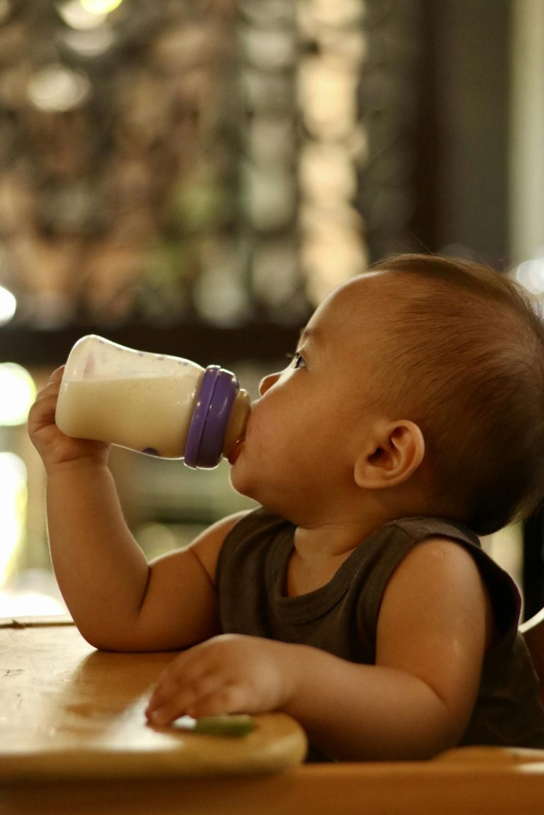 A cute baby enjoying milk from a bottle while sitting indoors. Warm lighting enhances the cozy ambiance.