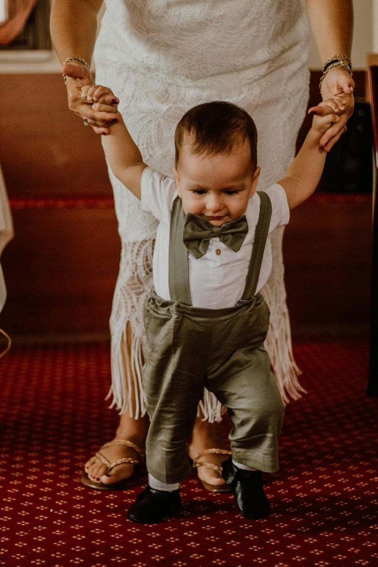 Cute toddler in bow tie and suspenders learning to walk with parent's help on a carpeted floor.